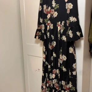 Svart blommig långklänning - Säljer en elegant svart långklänning med vackert blommönster i vitt och rosa. Klänningen har trekvartsärmar med volangdetaljer och en lös passform som ger en romantisk känsla. Perfekt för vår- och sommarfester!
