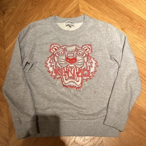 Grå sweatshirt från Kenzo - Säljer en grå sweatshirt från Kenzo med ett coolt tigertryck i rött och vitt. Tröjan är i bra skick men en liten röd fläck under märket och har långa ärmar. Perfekt för en avslappnad stil eller att bära till skolan. Superbekväm och stilren! Passar en 11-13 åring (själv på bilden är jag 13)