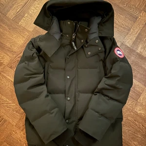 Canada Goose Wyndham - Säljer en svart dunjacka från Canada Goose i mycket bra skick. Jackan har en quiltad design med långärmade ärmar och en praktisk huva. Perfekt för kalla vinterdagar. Den har både dragkedja och knappar för extra skydd mot kyla. Märkets ikoniska logga syns på ärmen. Passar perfekt för vinterns alla äventyr! ❄️