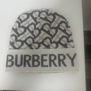 Burberry mössa - Snygg och stilren mössa från Burberry i grått och vitt med ett coolt mönster och stor logga framtill. Perfekt för att hålla sig varm och trendig under kyliga dagar. Passar både till vardags och lite finare tillfällen. One size