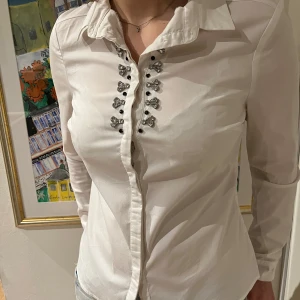 Vit skjorta med dekorativa knappar - Säljer en stilren vit skjorta med unika dekorativa knappar framtill. Skjortan har en klassisk krage och långa armar. 