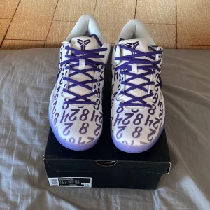 Kobe VIII 8 Protro sneakers i vitt och lila - Snygga Kobe VIII Protro sneakers i vitt med lila detaljer och unikt mönster. Skorna har en lila sula och snörning, samt en signaturdetalj på sidan. Perfekta för basketentusiaster.