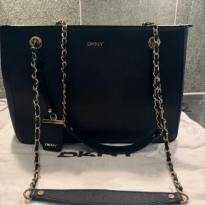 Svart axelväska från DKNY - Elegant svart axelväska från DKNY med guldfärgade kedjedetaljer och dragkedja upptill. Väskan har en stilren design med DKNY-logga framtill och är perfekt för en chic look.