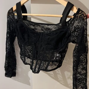 Svart spetskorsett - Snygg svart korsett i spets med långa ärmar och axelband. Perfekt för en kväll ute eller en speciell tillställning. Den har en elegant och sofistikerad design som framhäver figuren.