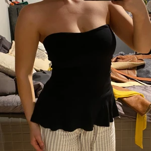 Svart axelbandslös topp - Snygg svart axelbandslös topp med volangdetalj i nederkant. Perfekt för en stilren och elegant look. Passar bra till både jeans och kjol.