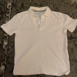 Vit kortärmad pikétröja från Hugo Boss - Säljer en stilren vit pikétröja från Boss med broderad logga på bröstet. Tröjan har en klassisk krage med knappar och är perfekt för en avslappnad men ändå uppklädd look.