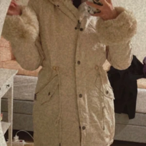 Beige parkajacka med pälsdetaljer - Säljer en mysig beige parkajacka med pälsdetaljer på ärmsluten och huvan. Jackan har knappar och dragkedja framtill samt praktiska fickor. Perfekt för kyliga dagar 🙂