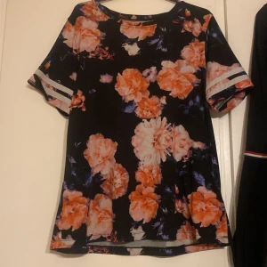 Blommig topp med korta ärmar - Säljer en svart topp med stora blommor i orange och rosa nyanser. Toppen har korta ärmar med randiga detaljer och en rund halsringning. Perfekt för en färgglad look!