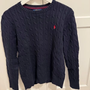 Mörkblå kabelstickad tröja från Ralph Lauren - Säljer en stilren mörkblå kabelstickad tröja från Ralph Lauren. Tröjan har en klassisk design med rund hals och det ikoniska röda logotypbroderiet på bröstet. Perfekt till exempelvis ett par jeans, pris kan diskuteras! Bara att höra av dig vid några funderingar!🤩🤩