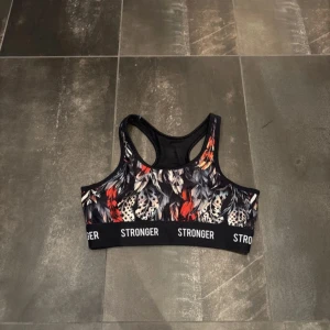 Sport bh - Snygg sporttopp från Stronger med ett färgstarkt mönster i svart, rött och vitt. Toppen har en brottarrygg och elastisk kant med logotyp. Perfekt för träning och aktivitet.