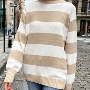 Beige och vit randig stickad tröja - Säljer en mysig stickad tröja i beige och vitt med breda ränder. Tröjan har en avslappnad passform och långa ärmar, perfekt för kyligare dagar. Den ribbade halsringningen och muddarna ger en klassisk look. En liten fläck finns som knappast syns