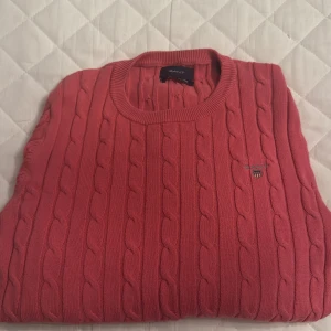 Rosa stickad tröja från Gant - Snygg rosa stickad tröja från Gant med kabelstickat mönster. Tröjan har rund hals och långa ärmar, perfekt för kyligare dagar. Den klassiska designen gör den lätt att matcha med olika outfits.