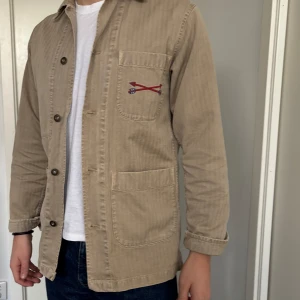 Beige overshirt med broderi - Säljer en beige overshirt från Chesapeake's med coolt broderi på ryggen. Jackan har knappar framtill och en bröstficka med broderad detalj. Perfekt för en avslappnad stil. Nypris 3,000