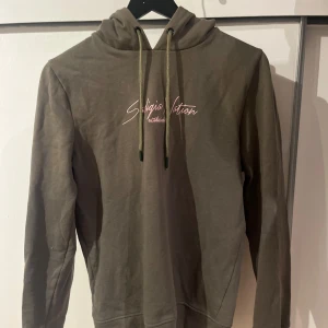 Olivgrön hoodie med tryck - Säljer en olivgrön hoodie med tryck på bröstet. Den har en justerbar huva med snören och långa ärmar. Perfekt för en avslappnad stil.