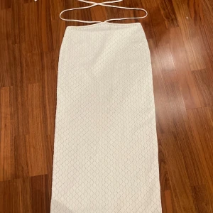 Vit lång kjol med snörning i midjan - Elegant vit lång kjol med snörning runt midjan. Kjolen har ett diskret rutmönster och är perfekt för sommarens festligheter. Den är tillverkad i ett lätt och bekvämt material.