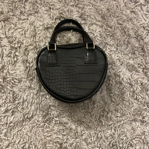 Svart handväska med krokodilmönster - Snygg svart handväska i syntet med krokodilmönster. Väskan har en unik form och kommer med ett avtagbart axelband. Perfekt för att ge din outfit en cool touch. Använd en gång💗