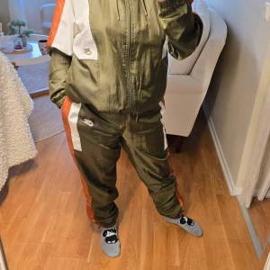 Träningsset - Snygg träningsjacka från Nike i grönt med orange och vita detaljer. Jackan har dragkedja och luva, perfekt för sportiga aktiviteter. Matchande byxor finns också tillgängliga. Det passar storlek M också (översize)