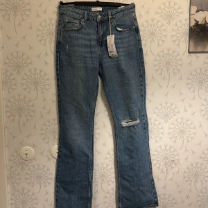 Blå jeans med slitningar - Snygga blå jeans med slitningar på ena benet. De har en klassisk femficksdesign och en knappgylf. Perfekta för en avslappnad stil.