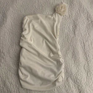 Vit axelbandslös topp - Elegant vit axelbandslös topp med en vacker blomdetalj på axeln. Perfekt för speciella tillfällen med sin stilrena design och mjuka material. Tror den är i storlek S. 