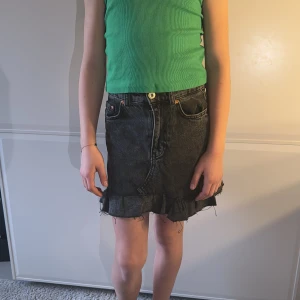 Svart jeanskjol med volang - Trendig svart jeanskjol med volangdetalj vid fållen. Kjolen har en klassisk femficksdesign och stängs med knapp och dragkedja framtill. Perfekt för en avslappnad stil.