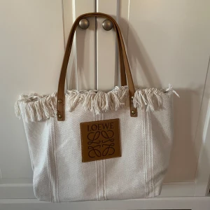 Vit shoppingväska från Loewe - Snygg vit shoppingväska från Loewe med fransdetaljer och bruna handtag. Väskan har en stor Loewe-logga framtill och är perfekt för att bära med sig allt man behöver. Den är rymlig och stilren, vilket gör den till ett utmärkt val för både vardag och speciella tillfällen.