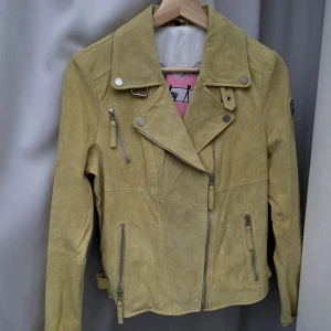 Gul mockajacka från Freaky Nation - Snygg gul mockajacka från Freaky Nation med coola dragkedjedetaljer och knappar. Jackan har en klassisk bikerstil med asymmetrisk dragkedja och flera fickor. Perfekt för att ge din outfit en edgy touch.