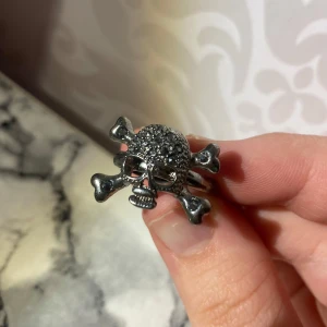 Silverfärgad dödskallering - Cool ring i form av en dödskalle med korslagda benknotor. Den är silverfärgad och har glittriga detaljer på skallen som ger en extra edge. Perfekt accessoar för att ge din stil en häftig touch. Den är även justerbar.