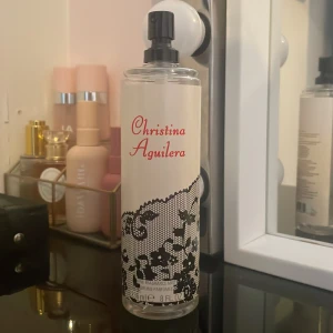 Christina Aguilera parfym - Elegant parfymflaska med svart spetsmönster och röd text. Flaskan har en praktisk sprayfunktion och rymmer 8 fl oz. Perfekt för den som vill ha en doft med stil och elegans.