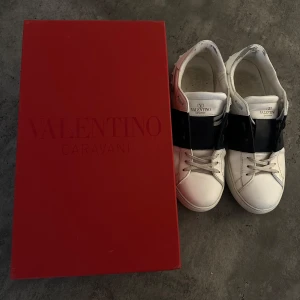 Valentino Garavani sneakers - Snygga vita Valentino Garavani sneakers med en bred svart rem över vristen. Skorna har klassisk snörning och kommer i originalförpackning. Perfekta för en stilren look.