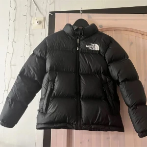 Svart dunjacka från The North Face - Säljer en svart dunjacka från The North Face. Jackan har en pufferdesign med dragkedja och hög krage för extra värme. Perfekt för kalla dagar. Väldigt bekväm och är i mycket bra skick. L i barnstorlek men passar xs i vuxenstorlek❤️❤️