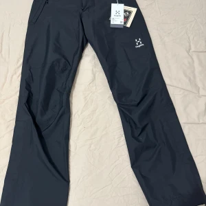 Svarta byxor från Haglöfs - Säljer ett par svarta byxor från Haglöfs. De har en rak passform och är tillverkade i ett syntetmaterial. Byxorna har en dragkedja och knappar framtill samt en diskret logga på benet.