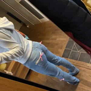 Blå jeans med rosa detaljer - Snygga blå jeans med unika rosa detaljer och spets på bakfickorna. De har en avslappnad passform med slitna partier och söta rosetter vid knäna. Perfekta för en trendig och avslappnad stil. Har själv sytt om från skinny till bootcut och även sytt på detaljerna, det är en av mina första jeans att sy på detaljer på så jag testade massor av olika saker som några blev bra och några blev mindre bra så därför säljs de för 150 kr❤️ (Det står M i jeansen men har sytt ner midjan å sytt in benen så mer S) 