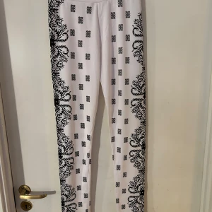 Vita leggings med svart mönster - Snygga vita leggings med ett detaljerat svart mönster längs sidorna och små mönster över hela byxan. Dekorerade med glittriga detaljer som ger en extra touch. Perfekta för att sticka ut i mängden!