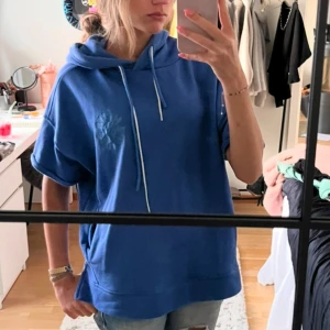 Blå hoodie med korta ärmar - Säljer en blå hoodie med korta ärmar och justerbar huva. Perfekt för en avslappnad stil. Den har en diskret logga på bröstet och en bekväm passform.