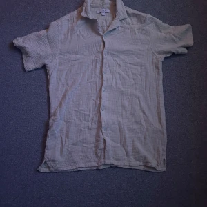 Beige New Yorker skjorta - Säljer en beige kortärmad skjorta från New Yorker i storlek S. Skjortan har en avslappnad passform och är tillverkad i ett lätt och luftigt material, perfekt för varma dagar. Den har knappar framtill och en klassisk krage.