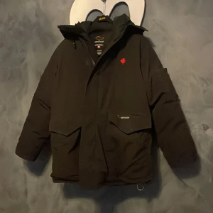 Svart dunjacka från Canada Goose - Säljer en svart dunjacka från Canada Goose med en stilren design och praktiska fickor. Jackan har en broderad röd logotyp på bröstet och dragkedja framtill. Perfekt för kalla vinterdagar.