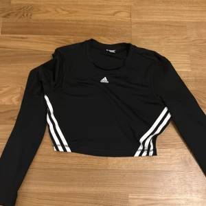 Svart croppad topp från Adidas - Snygg svart croppad topp från Adidas med vita ränder längs sidorna. Perfekt för en sportig look. Toppen har långa ärmar och en rund halsringning.