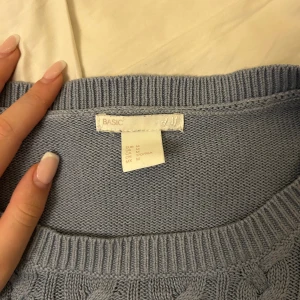 Blå stickad tröja från H&M - Säljer en mysig grå stickad tröja från H&M med flätmönster. Tröjan har långa ärmar och en rund halsringning. 