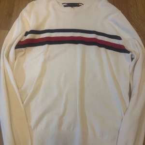 Vit tröja med randigt mönster från Tommy Hilfiger - Snygg vit tröja från Tommy Hilfiger med randigt mönster i marinblått och rött över bröstet. Tröjan har långa ärmar och en rund halsringning. Perfekt för en stilren look. Passar mellan s-m