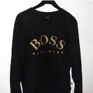 hugo boss tröja - Snygg svart tröja från Hugo Boss med guldtryck på bröstet. Tröjan har långa ärmar och en klassisk passform. Perfekt för en stilren look.