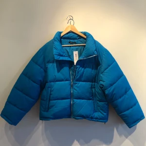 Blå pufferjacka - Säljer en oanvänd & helt ny blå pufferjacka. Jackan har en dragkedja framtill & silverdetaljer. Den är i storlek XL men passar även mindre för en oversized look. Jag på bilden har vanligtvis S/M . Perfekt för både höst och vinter!
