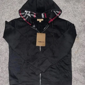 Burberry zip hoodie - En Burberry hoodie är en stilren och bekväm plagg, ofta med varumärkets ikoniska rutmönster eller logotyp. Den är tillverkad av högkvalitativa material och har en dragsko på huvan samt fickor, perfekt för avslappnad användning.