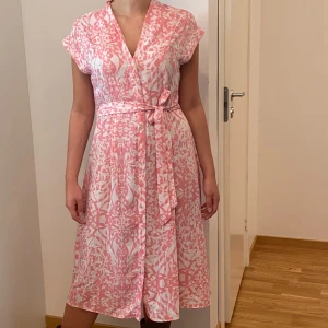Rosa och vit omlottklänning - Säljer en superfin rosa och vit omlottklänning med ett abstrakt mönster. Klänningen har korta ärmar och ett knytband i midjan för en smickrande passform. Perfekt för sommarens alla tillfällen! 🌸