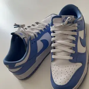 Säljer ett par Nike Dunk sneakers i en snygg blå och vit färgkombination. Väldigt nya, användes bara två gånger, köpte de för 1600kr. Skorna har klassisk snörning och är i skinnmaterial. Perfekta för både vardag och träning!