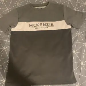 Säljer en stilren grå t-shirt från McKenzie. Den har en vit rand över bröstet med McKenzie-loggan tryckt i grått. Perfekt för en avslappnad stil eller träning. T-shirten är i perfekt skick och passar perfekt för vardagsbruk.