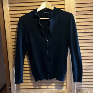 Svart kofta från Hugo Boss - Säljer en stilren svart kofta från Hugo Boss, tillverkad av finaste italienska garnet. perfekt för både höst och vinter. Koftan är 9/10 i skick och har en broderad logga på bröstet. Storlek M men krympt i tvätten så passar XS