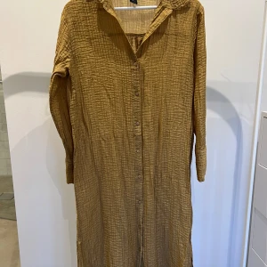 Beige långklänning från Lindex -  långklänning från Lindex. Den har en avslappnad stil med långa ärmar och knappar hela vägen ner. Perfekt för både vardag och lite finare tillfällen. Materialet känns lätt och luftigt, vilket gör den perfekt för vår och sommar!