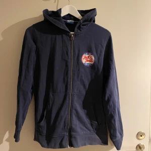 Blå huvtröja med dragkedja - Säljer en snygg blå huvtröja från Jack & Jones. Den har en dragkedja framtill och en cool patch på bröstet. Perfekt för en avslappnad stil och passar bra till både jeans och joggers. Tröjan har långa ärmar och en skön huva. Medium