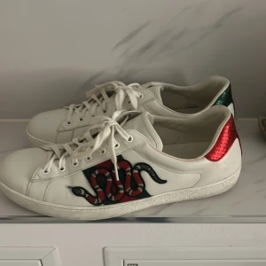 Gucci sneakers med ormmönster - Säljer ett par snygga vita Gucci sneakers med ett coolt ormmönster på sidan. Skorna har röda och gröna detaljer på hälen och klassiska snörning framtill. Perfekta för att ge din outfit en lyxig touch!