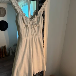 Vit klänning med volangdetaljer - Säljer en superfin vit klänning med volangdetaljer på axelbanden. Den har en söt knytning framtill och är perfekt för sommardagar. Klänningen är i ett mjukt och bekvämt material, vilket gör den idealisk för både vardag och fest. något kortare modell! 